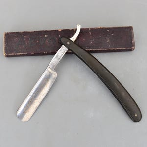 Könnte beinhalten: Ein offenes Rasiermesser mit schwarzem Griff und silberner Klinge, auf einer grauen Oberfläche. Das Rasiermesser liegt neben einer braunen rechteckigen Schachtel mit verblichenem Text. Die Klinge hat die Nummer "16".