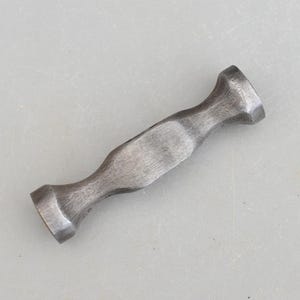 May include: A small, hand-forged metal hammer with a dark, weathered finish. The hammer has a rounded head on each end and a slightly curved handle. The metal has visible tool marks, giving it a rustic appearance. The hammer is designed for precision work.