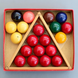 May include: A vintage billiard ball set in a red box. The set includes a wooden triangle rack and 15 colourful balls: red, yellow, black, pink, brown, blue, and green. The balls are arranged in a triangle formation inside the box.