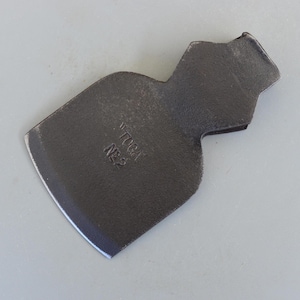 以下が含まれることがあります： 湾曲した刃と長方形の上面を持つ、濃い灰色の金属製のハチェットヘッド。「TOGA NE2」の文字が側面に刻印されています。ハチェットヘッドは、明るい灰色の表面にあります。