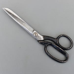 Puede incluir: Tijeras de metal vintage con una hoja plateada y mangos negros. Las tijeras están abiertas, mostrando las afiladas hojas. Los mangos tienen orificios para los dedos. Estas tijeras probablemente se utilizan para cortar tela o papel. Las hojas miden unos 20 cm.