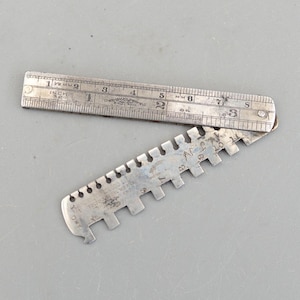May include: A vintage, silver-toned metal gauge with a hinged ruler. The ruler is marked with inches and millimeters. The gauge has a series of notched teeth and is marked with numbers and text. The tool is likely used for measuring or checking dimensions.