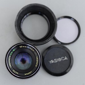 Peut inclure: Un objectif d'appareil photo Yashinon DS-M 50mm f/1.7 à mise au point automatique avec un pare-soleil noir, un bouchon d'objectif et un filtre transparent. Le bouchon d'objectif porte le mot "YASHICA" en relief. L'objectif est marqué "JAPAN".