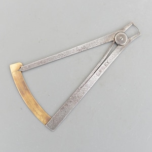Puede incluir: Un calibrador de metal vintage con un extremo curvo de latón y un cuerpo plateado. El calibrador está abierto, mostrando la escala de medición. El nombre "C. RILEY" está grabado en uno de los brazos. La herramienta está sobre un fondo gris.