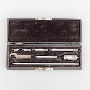May include: A grey rectangular case, open to reveal a dark brown velvet interior. Inside are several silver metal tools, including rods, a small wrench, and a measuring device. The tools are neatly arranged within the case.