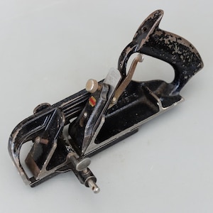 May include: A vintage black and silver hand plane with a curved handle. The tool has a blade, adjustment knobs, and a lever cap. The metal shows signs of wear, indicating its age and use. A classic woodworking tool.