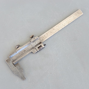 Pode incluir: Um paquímetro de metal prateado com uma régua na parte superior. O paquímetro tem uma mandíbula deslizante e uma mandíbula fixa. A régua é marcada em polegadas.