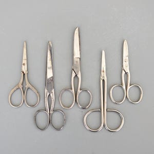 May include: Five silver scissors of varying sizes and styles. The scissors are arranged in a row on a light grey background.