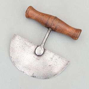 Puede incluir: Un cuchillo mezzaluna vintage con una hoja curva de acero inoxidable y un mango de madera. El mango es de color marrón oscuro y tiene forma redondeada. La hoja muestra signos de desgaste. Diseñado para picar hierbas o verduras.