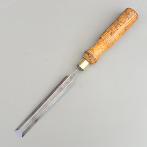 Könnte beinhalten: Ein Holzbeitel mit einem hellbraunen Holzgriff mit dunklen Flecken und einer Messingzwinge. Die silberne Metallklinge ist abgeschrägt und für präzise Schnitte in Holz konzipiert. Das Werkzeug ist etwa 20 cm lang.