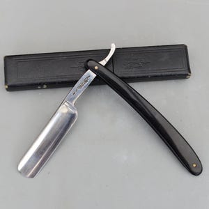 Puede incluir: Una navaja de afeitar con una hoja plateada brillante y un mango negro. La navaja está abierta, junto a un estuche negro. La hoja tiene texto. Es una herramienta de aseo.