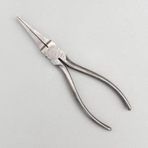 May include: A pair of long-nose pliers, a hand tool with pointed jaws for gripping and bending. The pliers are made of metal and have a silver finish. The tool has a pivot point and curved handles for leverage. The tool has text on the side.