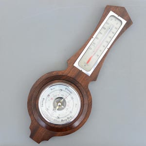 Puede incluir: Un barómetro y termómetro de madera vintage. El barómetro tiene una esfera circular con lecturas de presión, mientras que el termómetro muestra temperaturas en grados Fahrenheit. El marco de madera tiene un acabado oscuro. La temperatura es de unos 20 grados Celsius.