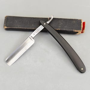 Könnte beinhalten: Ein Vintage-Rasiermesser mit schwarzem Griff und silberner Klinge. Das Rasiermesser ist geöffnet und zeigt die scharfe Kante. Die Klinge ist mit Text markiert. Eine schwarze Hülle befindet sich im Hintergrund.