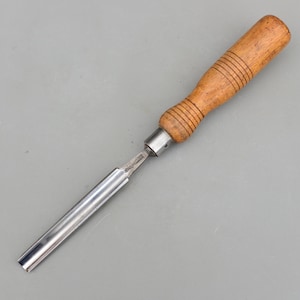 以下が含まれることがあります： 木工用のノミ。明るい茶色の木製ハンドルと銀色の金属刃が付いています。このツールは木材を成形するために設計されています。ハンドルは約10cmの長さです。