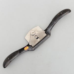 May include: A black and silver hand plane with a curved handle and a blade. The handle has a textured grip. The tool has a metal blade and a red and yellow sticker that says "STANLEY".