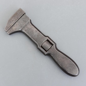 May include: A dark grey vintage adjustable wrench. The wrench has a textured adjustment knob and a tapered handle. The head of the wrench has a wide opening. This tool is designed for tightening or loosening nuts and bolts. The wrench is made of metal.
