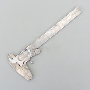 May include: A vintage silver caliper tool with inch and mm measurements. The tool has a sliding mechanism and a pointed jaw for precise measurements. The caliper is made of metal and is set against a grey background.