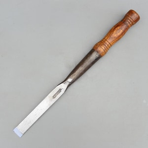 May include: A wood chisel featuring a long, flat, silver-colored metal blade with a sharp, beveled edge and a dark, polished wooden handle with decorative grooves. The tool has a tapered metal bolster connecting the blade to the handle.