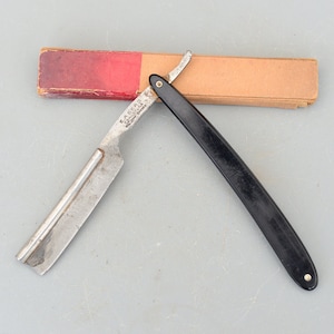 May include: A vintage straight razor with a black plastic handle and a silver blade. The razor is in a cardboard box with a red and brown design.