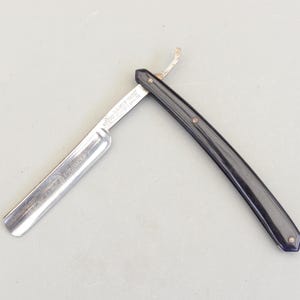 May include: A straight razor with a black handle and a silver blade, lying open. The blade has etched text. The handle is attached to the blade with a hinge. The razor is on a light grey surface.