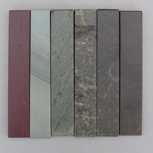 May include: Six rectangular sharpening stones arranged side-by-side in a row. The stones feature varied textures and colors, including deep maroon, light gray, olive green with diagonal streaks, mottled dark gray, charcoal, and solid dark brown.