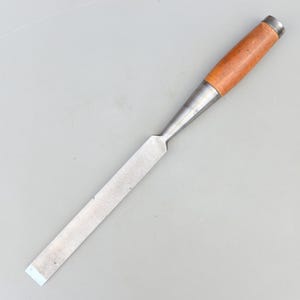 May include: A woodworking chisel with a wooden handle and a metal blade. The handle is light brown with a silver ferrule. The blade is rectangular and silver. The tool is designed for shaping wood.