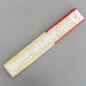 Könnte beinhalten: Ein Vintage-Rechenschieber mit cremefarbenem Gehäuse und roten Kanten. Der Rechenschieber verfügt über mehrere Skalen und Markierungen für mathematische Berechnungen. Das Gerät ist für die schnelle und genaue Durchführung komplexer Berechnungen konzipiert.