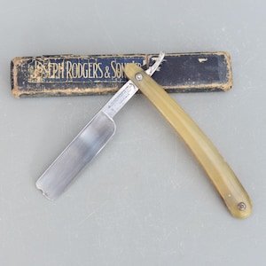 Könnte beinhalten: Ein Vintage-Rasiermesser mit einem Knochengriff und einer silbernen Klinge. Das Rasiermesser befindet sich in seiner Originalverpackung, die schwarz ist mit goldenen Lettern, die "Joseph Rodgers & Sons Ltd." lesen.