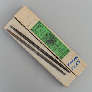 Op de afbeelding: Twee kleine metalen vijlen in hun originele verpakking. De dozen zijn wit met blauwe letters die "SMALL FILES" lezen. De grotere doos heeft een groen label met de tekst "NICHOLSON FILE CO. WARRANTED THE BEST FILES MADE IN THE WORLD."
