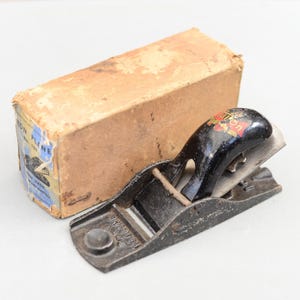 Könnte beinhalten: Ein vintage Handhobel aus schwarzem Metall mit einem Holzgriff. Der Hobel befindet sich in seiner Originalverpackung, die mit "Union Plane No. 100" und "Ten Hand Warranted" beschriftet ist.