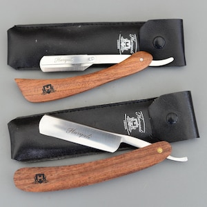 Może przedstawiać: Dwie brzytwy z drewnianymi uchwytami i ostrzami ze stali nierdzewnej, każda w czarnym skórzanym etui. Ostrza są grawerowane napisem "Haryali". Uchwyty są w kolorze czerwonobrązowym, a etui mają zapięcie na zatrzask.