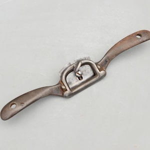 Puede incluir: Un cuchillo de tracción vintage con una hoja curva y dos mangos. La herramienta está hecha de metal con un aspecto oxidado y envejecido. Los mangos tienen agujeros en los extremos. Esta herramienta para trabajar la madera está diseñada para dar forma a la madera.