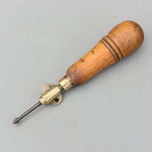May include: A vintage awl with a wooden handle and a metal tip. The handle is light brown with dark brown rings. The metal tip is dark gray and has a pointed end. The awl has a brass fitting.
