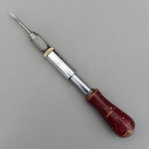 Puede incluir: Un destornillador de cabeza plana vintage con un eje de metal plateado y un mango de madera rojo oscuro. El mango muestra signos de desgaste. El destornillador está diseñado para apretar o aflojar tornillos.