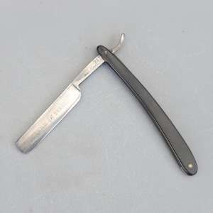 Könnte beinhalten: Ein Vintage-Rasiermesser mit schwarzem Kunststoffgriff und einer silbernen Klinge. Die Klinge ist mit dem Text "Hand Forged" beschriftet.
