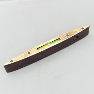 May include: A vintage spirit level with a dark wood body and brass accents. The level features a clear vial with a green liquid and is designed to measure the horizontal or vertical alignment of a surface. The tool is likely used for carpentry or construction.