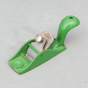 May include: A small, vintage green hand plane with a silver blade and a curved handle. The blade is marked with the word "KUNZ". This woodworking tool is designed for smoothing and shaping wood surfaces.