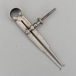 Può includere: Un calibro esterno argentato vintage con regolazione a vite. Lo strumento ha due gambe curve e un'impugnatura circolare. Il testo "THE S.S. STARRETT CO. ATHOL MASS. U.S.A." è visibile sul calibro.