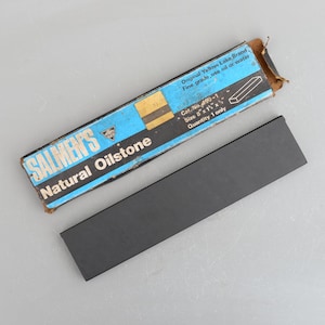 May include: A blue and white box with the text "SALMEN'S Natural Oilstone" and "Original Yellow Lake Brand Fine grade, use oil or water Cat. No. 490-1 Size 8"x 1 1/4"x 1/2" Quantity 1 only". The box is next to a black sharpening stone.