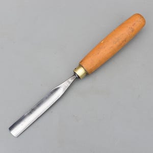 Könnte beinhalten: Ein Holzschnitzbeitel mit einer gebogenen, polierten Stahlklinge und einem hellbraunen Holzgriff. Der Griff ist zylindrisch und wirkt abgenutzt. Eine Messingzwinge verbindet die Klinge mit dem Griff. Das Werkzeug ist zum Schnitzen und Formen von Holz bestimmt.