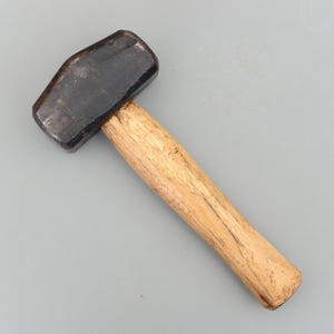 May include: A sledgehammer with a dark, rectangular head and a light brown wooden handle. The tool is designed for heavy-duty tasks. The hammer head is made of steel. The handle is made of wood.