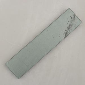 Peut inclure: Une pierre à aiguiser rectangulaire grise avec une surface légèrement texturée. La pierre est de couleur gris clair et présente quelques marques grises plus foncées à une extrémité. Elle est conçue pour affûter les couteaux et autres lames.