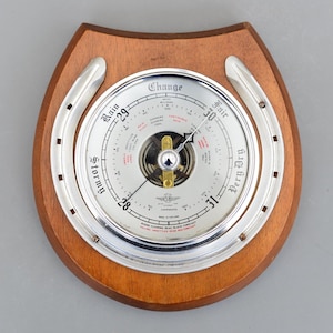 Puede incluir: Un barómetro vintage con esfera plateada y marco de madera en forma de herradura. El barómetro indica las condiciones meteorológicas. El marco tiene un acabado pulido. El diámetro es de unos 15 cm.