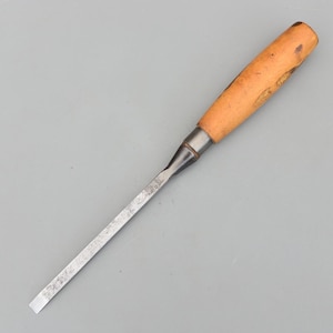 May include: A woodworking chisel with a light brown wooden handle and a silver metal blade. The handle has a cylindrical shape and is attached to the blade with a metal ferrule. The blade is rectangular and tapers to a flat edge.
