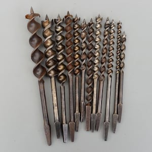 Może przedstawiać: Kolekcja antycznych wierteł świdrowych, każde ze spiralnym wzorem i spiczastą końcówką. Wiertła różnią się rozmiarem i są wykonane z metalu, o zwietrzałym, postarzanym wyglądzie. Narzędzia są ułożone na neutralnym tle.