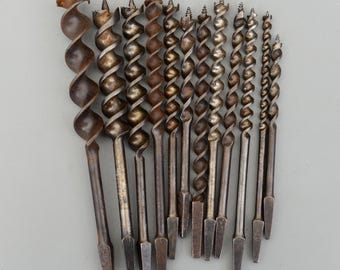 May include: A collection of antique auger drill bits, each with a spiral design and a pointed tip. The bits vary in size and are made of metal, with a weathered, aged appearance. The tools are arranged on a neutral background.