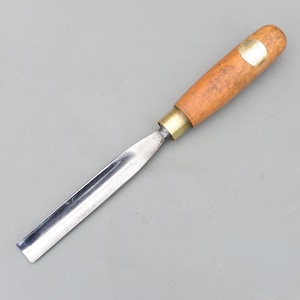 以下が含まれることがあります： 木工用のノミ。木製の柄と金属製の刃が付いています。柄は薄茶色で、真鍮のフェルールと小さな真鍮プレートが付いています。刃は銀色で角度が付いています。