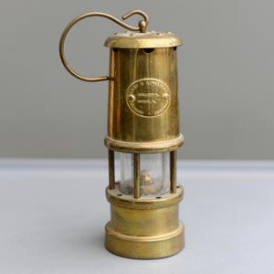 May include: A vintage brass miner's lamp with a handle and a glass enclosure. The lamp has a cylindrical shape with a round base and top. The lamp has a label that reads "LAMP & LIMELIGHT COMPANY, SERIAL No."
