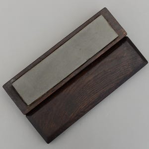 May include: A rectangular sharpening stone set in a dark brown wooden case. The stone is a light grey colour and is rectangular. The wooden case is open, revealing the stone. The wood grain is visible.